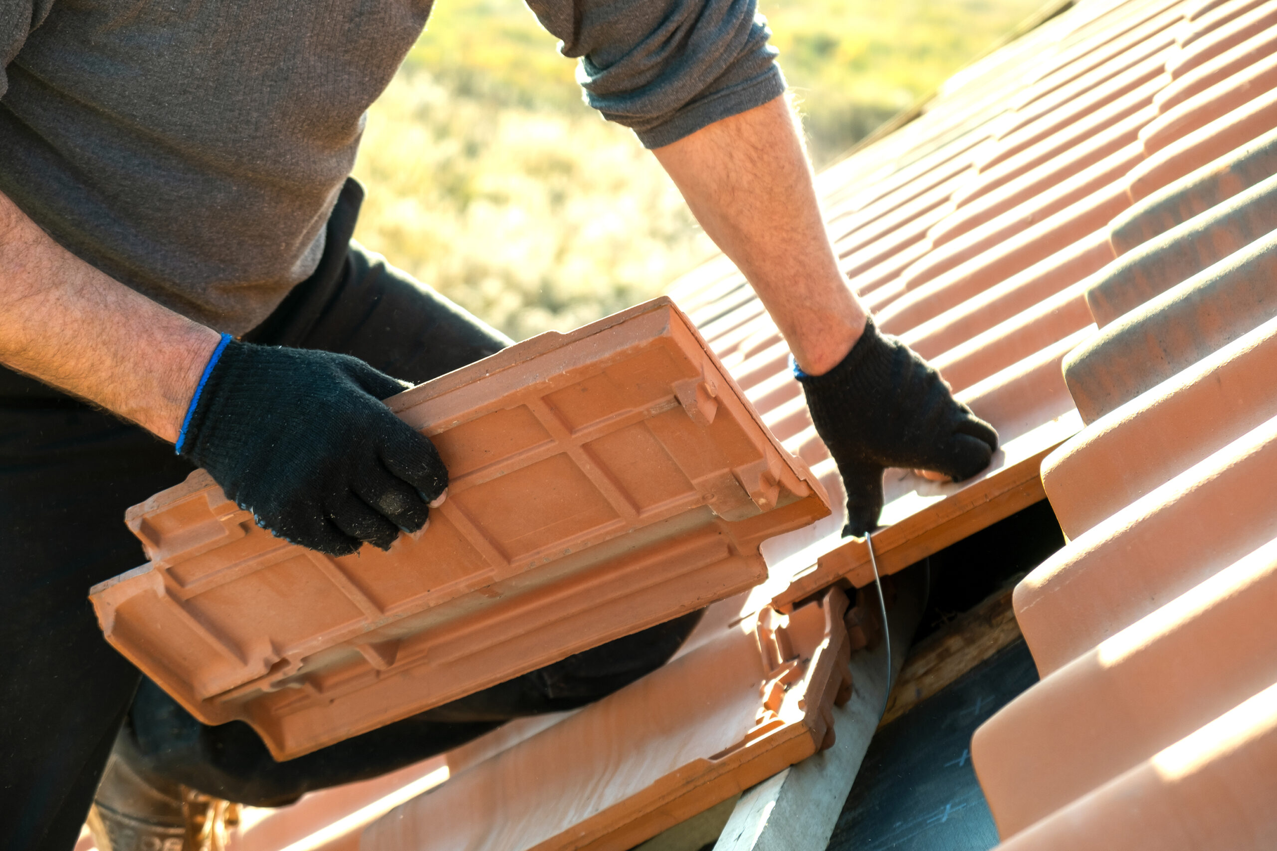 Closeup of worker hands installing yellow ceramic roofing tiles mounted on wooden boards covering residential building roof under construction.